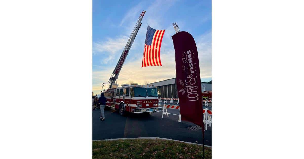 photo of Ayer fire department truck holding up an American flag with a Loaves & Fishes Food Pantry feather style flag in the foreground