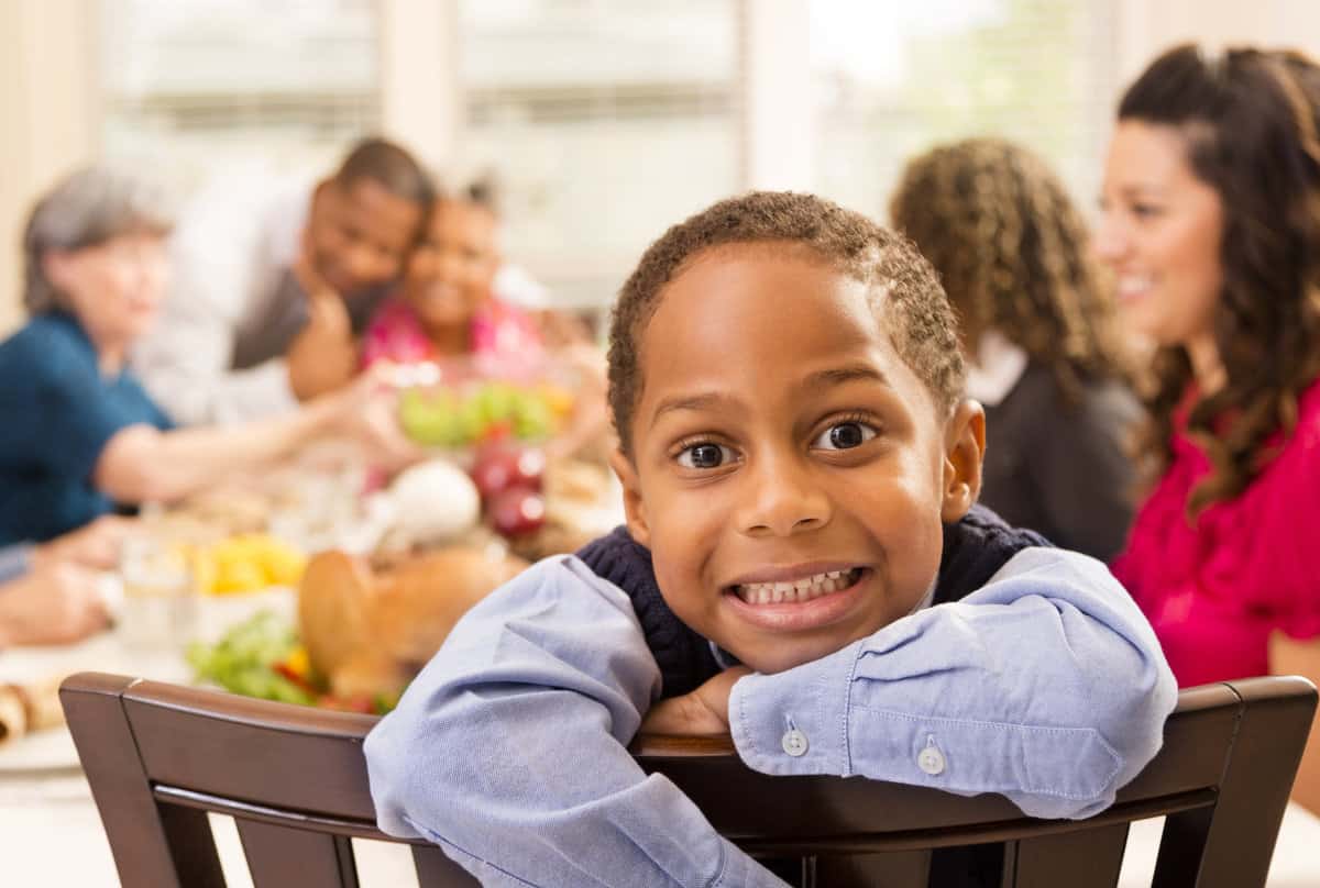 Multi-ethnic group of family and friends gather together for a holiday dinner.  Thanksgiving or Christmas season. Cute African descent little boy poses for the camera in the foreground.  Other family and friends sitting around dinner table in background.